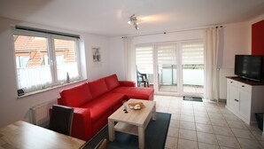 A living room with a red sofa, a wooden coffee table, a TV on a cabinet, and a view of the outside through large windows.