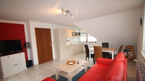 A modern living room with a red sofa, a wooden coffee table, and a flat-screen TV. There is a kitchen area with white cabinets and a dining table with chairs.