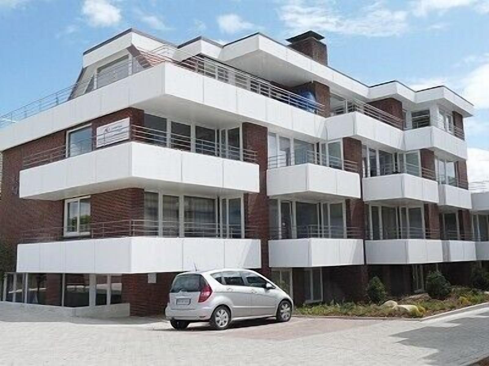 A modern multi-story building with balconies, large windows, and a car parked in front.