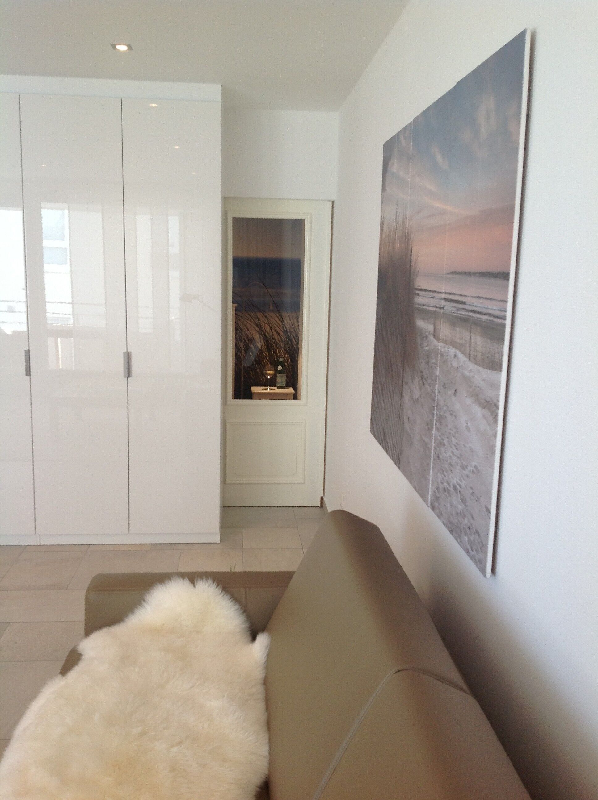 Una sala de estar con un sofá color beige, un tapete blanco y mullido, un cuadro grande con una playa y un armario blanco.