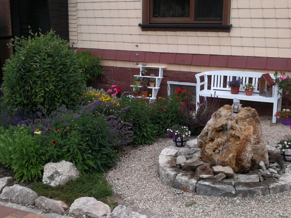 Un jardín con diversas plantas y flores, un banco blanco y un elemento decorativo de rocas.