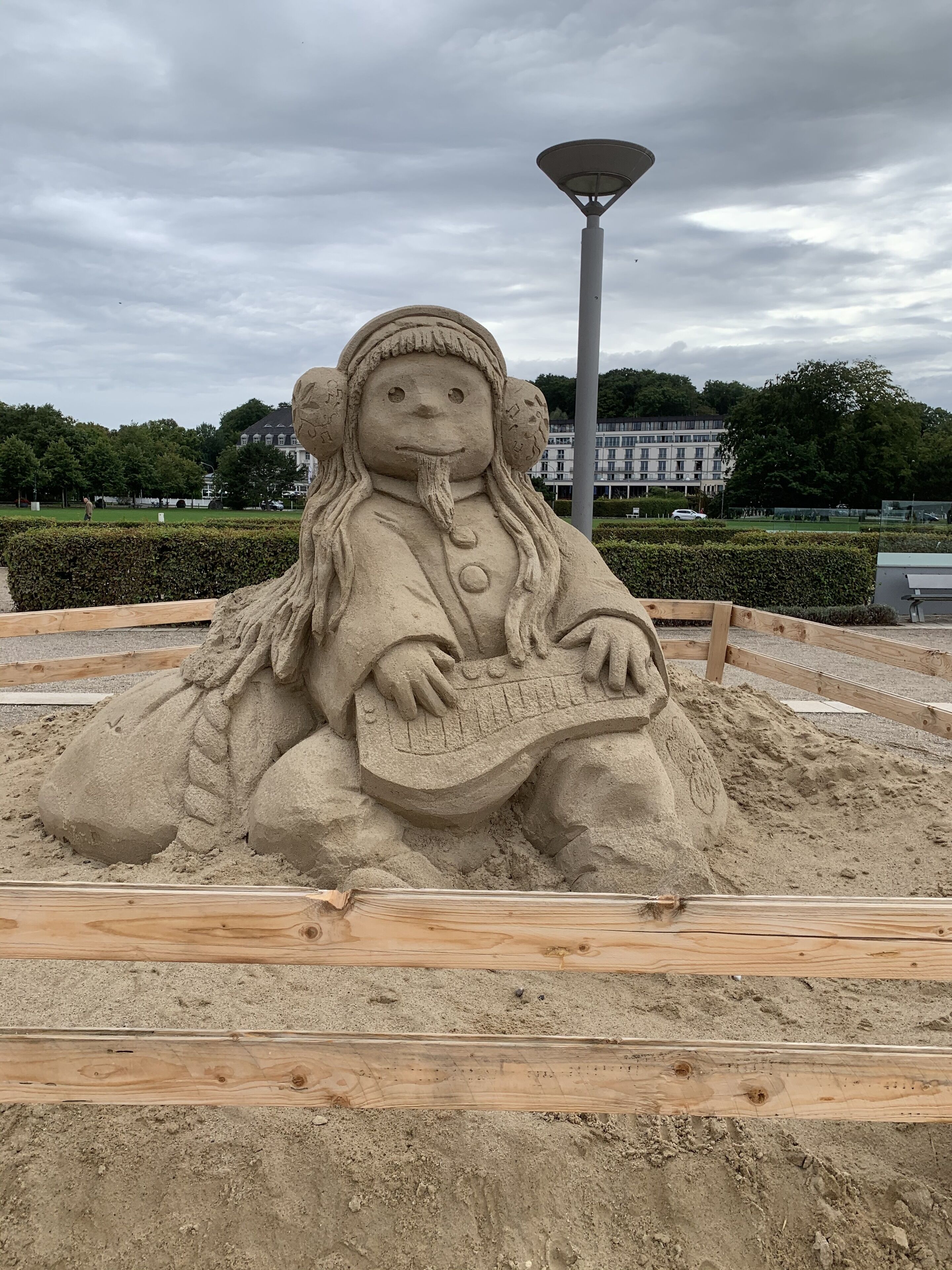 A sand sculpture of a figure with a keyboard, surrounded by sand and wooden planks, with a lamp post and buildings in the background.
