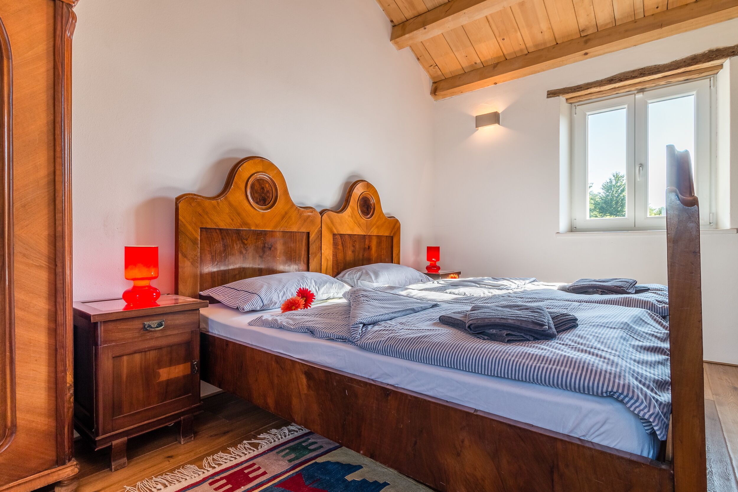 A wooden four-poster bed with striped bedding, a nightstand with a red lamp, and a window with a view of greenery.