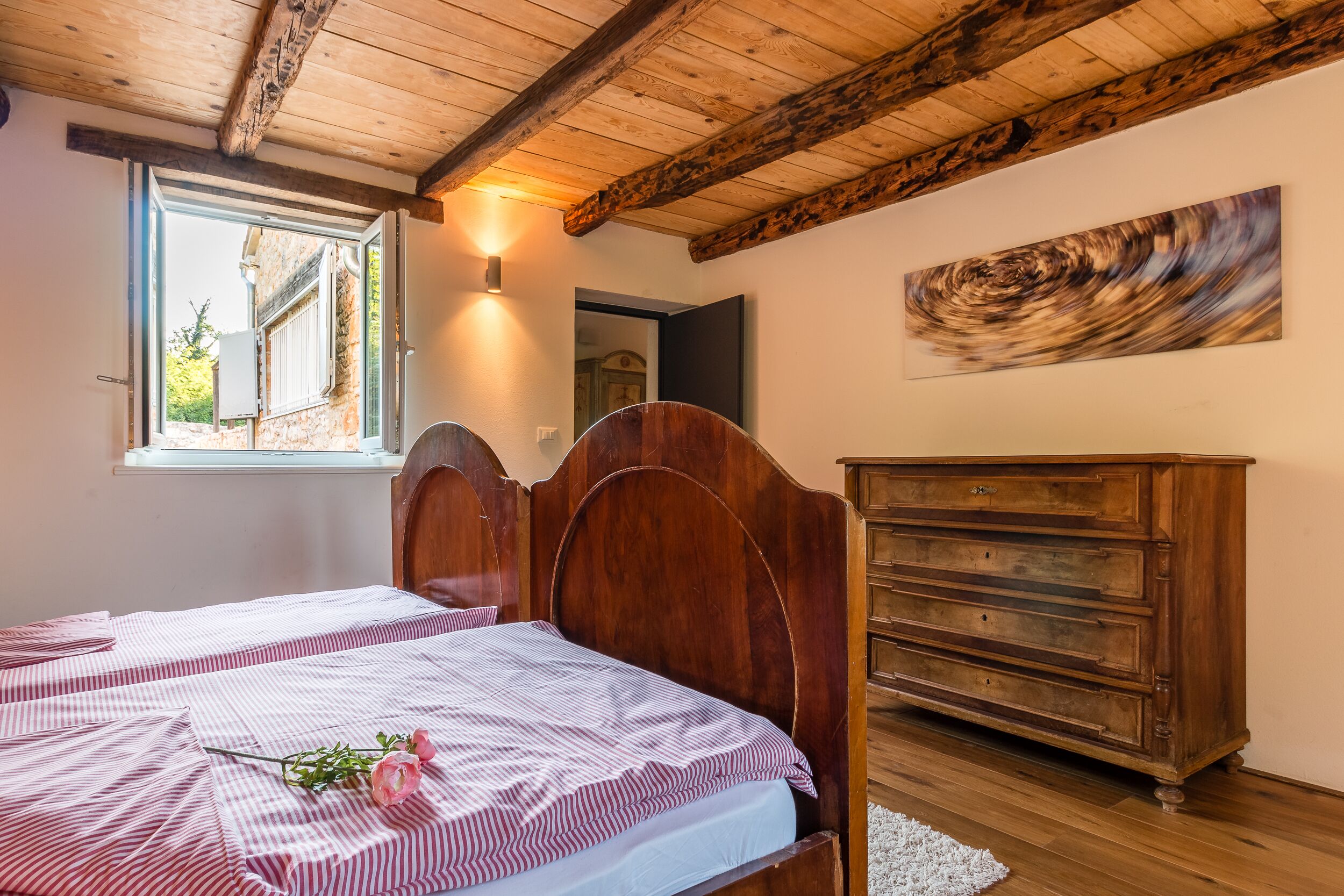 A bedroom with two beds, wooden headboards, a dresser, a window with a view, and a painting on the wall.