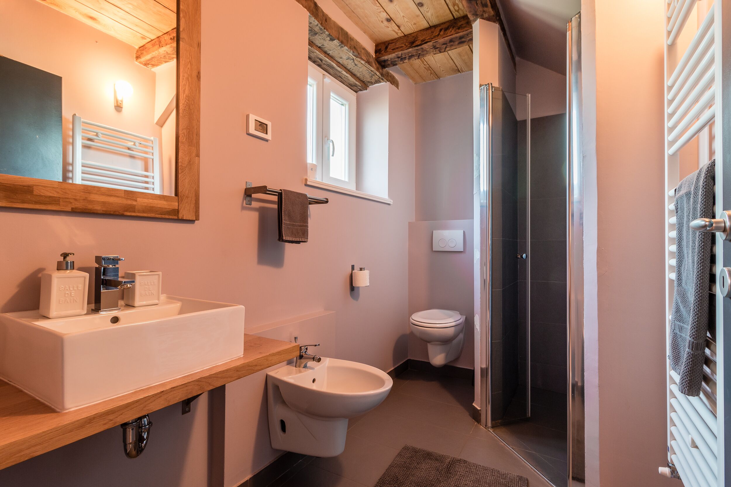 A bathroom with a wooden vanity, a white sink, a bidet, a toilet, and a glass shower enclosure.