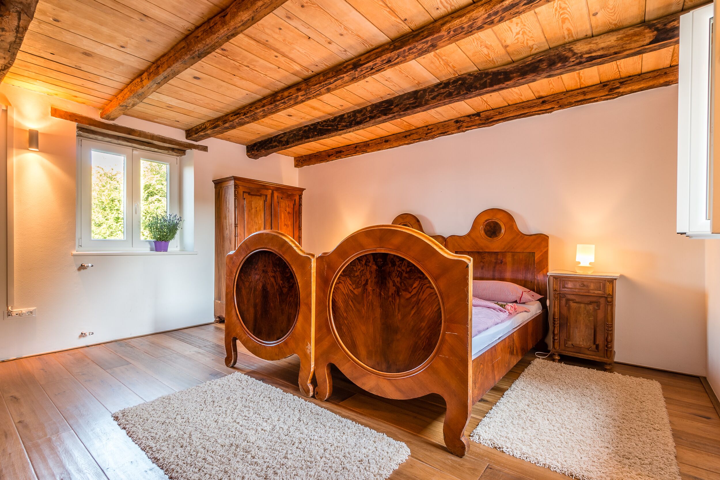 Ein Schlafzimmer mit Holzboden, einem Bett mit Holz Kopfteil, einem Nachttisch, einem Schrank und einem Fenster mit Blick auf Grünflächen.