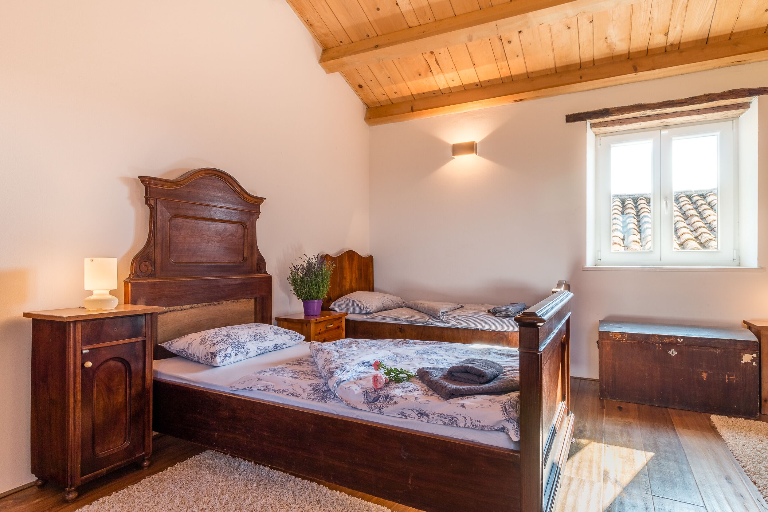 Une chambre à coucher avec un lit en bois, une table de chevet, une commode, une fenêtre donnant sur des immeubles et un plafond avec des poutres en bois.