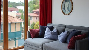 A grey sofa with multiple cushions, a framed artwork on the wall, and a view of buildings through a window with red curtains. - Sunny, east and south situated comfortable 4-room apartment. (Kühlungsborn)