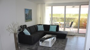 A modern living room with a sectional sofa, a coffee table, and a large window offering a view of the outdoors. - Wilhelmshaven Südstrand: Hochw. Duplex apartment right on South Beach (Wilhelmshaven)