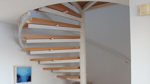 A spiral staircase with wooden steps and white railings, a small table, and a chair with a blue cushion in a room with grey tiled flooring. - Wilhelmshaven Südstrand: Hochw. Duplex apartment right on South Beach (Wilhelmshaven)