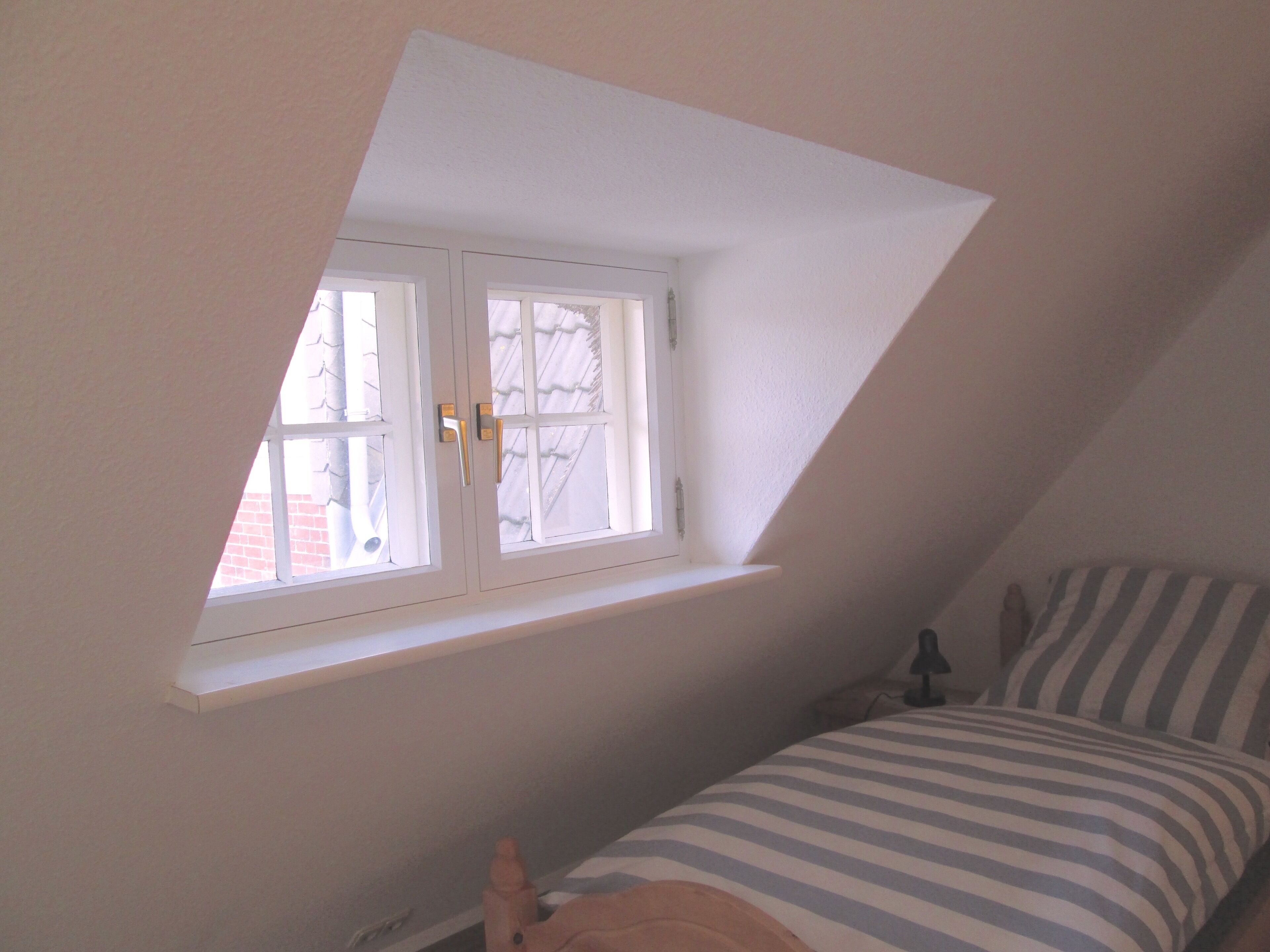 Une chambre avec un plafond en pente, une fenêtre à cadre blanc, un lit avec une literie à rayures et une table de chevet avec une lampe.