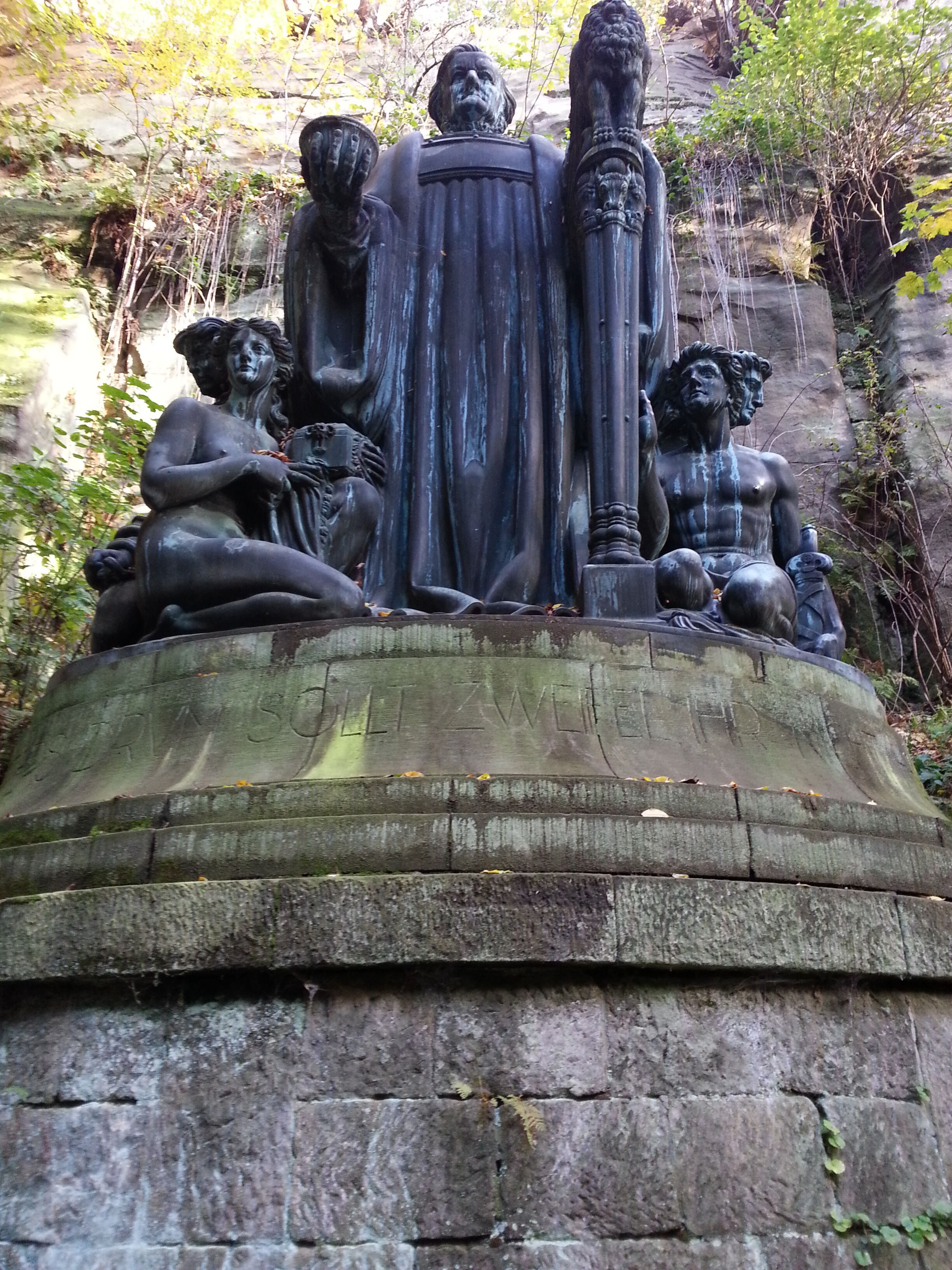 Un monument de pierre avec une figure centrale, flanquée de figures plus petites, le tout se détachant sur un décor naturel.