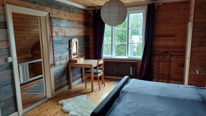 A wooden cabin room with a bed, a desk, a chair, a mirror, and a window with a view of trees.