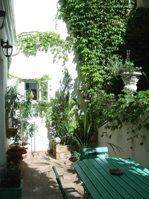 Un patio iluminado por el sol con un banco verde, plantas en macetas y una pared blanca cubierta de hiedra.