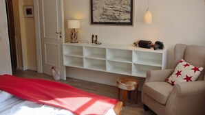 Una habitación con una cama, una silla con un cojín rojo en forma de estrella, un estante blanco con objetos decorativos, una obra de arte enmarcada y una puerta.