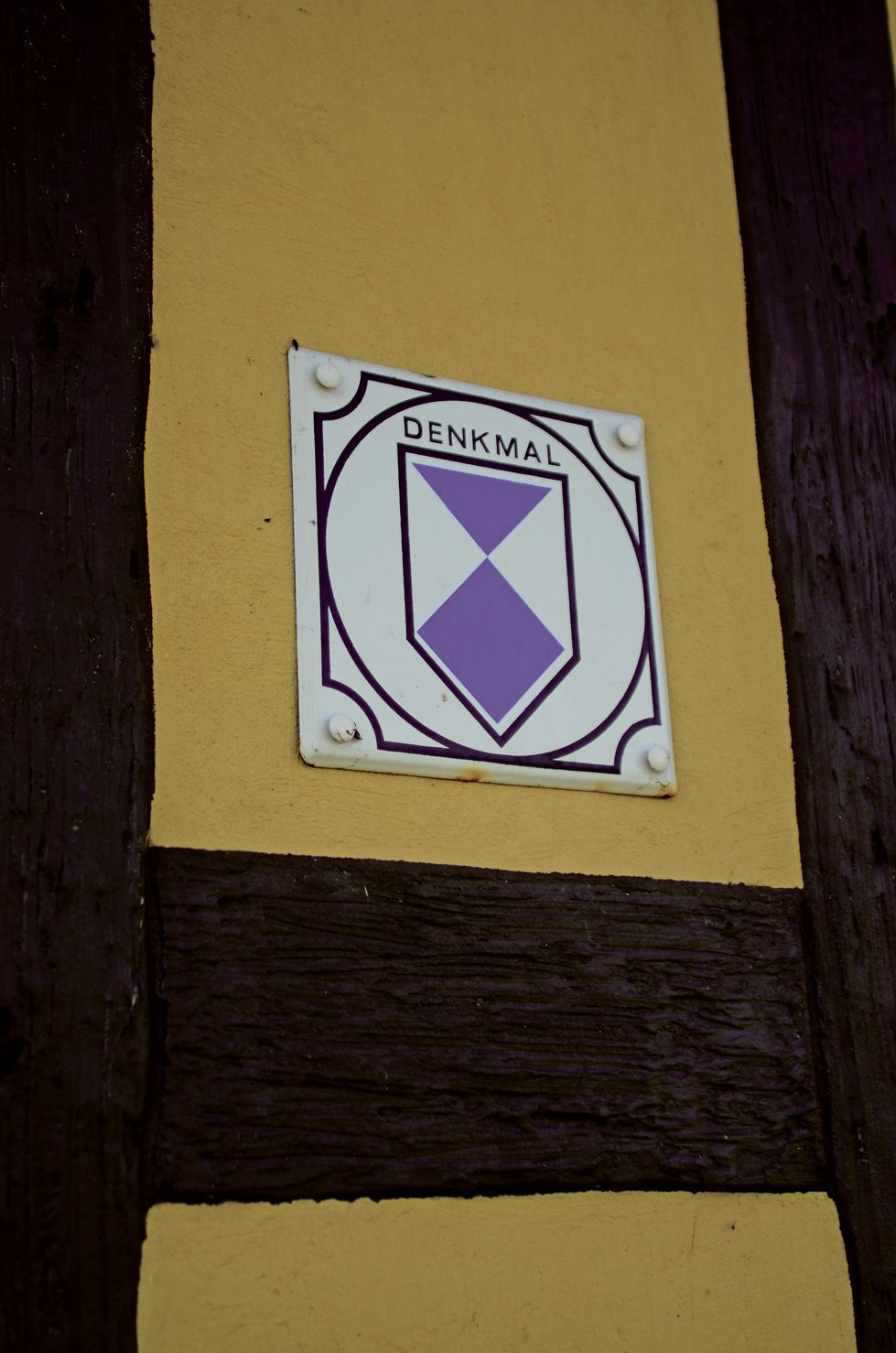 Ein Schild mit dem Wort 'DENKMAL' an einer gelben Wand mit dunklem Braunton im unteren Bereich.
