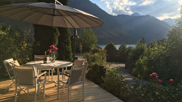 A wooden deck with a round table and chairs, an umbrella, and a vase with flowers. - Apartment with seaview, 2 bedrroms. own garden, 5 min. to lake (Tirol)