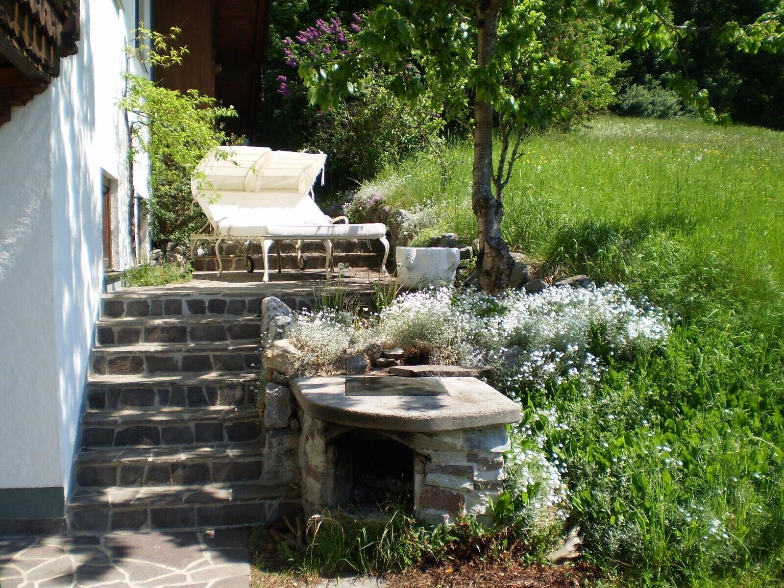 A stone staircase leading to a white door, a white outdoor chair, and a stone bench in a garden with white flowers and green grass.
