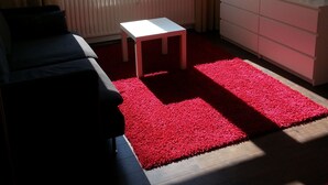 A living room with a sofa, a small white table, a TV on a white cabinet, and a red carpet.