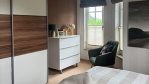 A bedroom with a bed, a green armchair, a dresser, and a window with a view of greenery. - Modern, spacious, high quality furnished, apartment for 2-3 people (Sehlen)