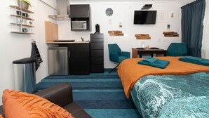 Une chambre d’hôtel moderne équipée d’une kitchenette, d’un lit recouvert d’une couverture orange, d’un canapé bleu et d’un téléviseur fixé au mur.