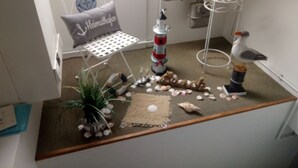 A small, cozy room with a nautical theme, including a small table with a lighthouse model, a chair with a cushion, and a decorative bird. -  Apartments directly on the North Sea in Schleswig-Holstein (Friedrichskoog)