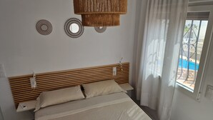 A bedroom with a wooden headboard, a bed with beige bedding, two wall-mounted decorative mirrors, and a view of a swimming pool through a window with sheer curtains. - HOLIDAY HOUSE DIRECTLY BY THE SEA (Oliva Playa)