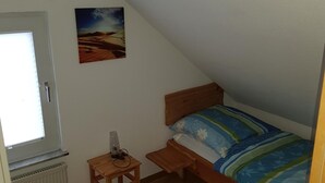 Una stanza piccola e con il soffitto spiovente, con un letto di legno, un tavolino, un termosifone e una finestra.