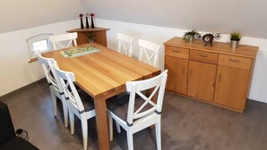 Zona pranzo con tavolo in legno e sedie bianche, un piccolo mobile con oggetti decorativi e una finestra con persiane.