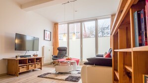 Ein Wohnzimmer mit Flachbildfernseher, einer Couch, einem Sessel, einem Couchtisch und einem Bücherregal.