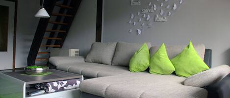 A modern living room with a grey sofa, green pillows, a white side table, and a wall decoration with butterflies and the words "Love sweet home".