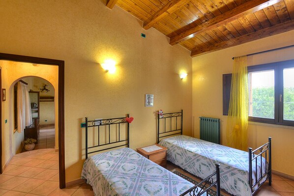 A bedroom with two beds, wooden ceiling beams, and a window with yellow curtains.