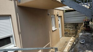 Ein schmaler Außenkorridor mit beiger Wand, Metalltreppe, Steinwand und einem kleinen Gartenbereich mit Blumentöpfen.