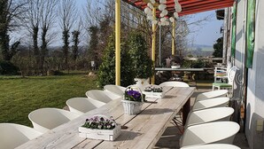 A covered outdoor dining area with a long wooden table and white chairs, a grill, and a view of the countryside. - Room in an old manor with an artistic atmosphere (neukalen)