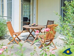 An outdoor seating area with a wooden table and chairs, a small table with a vase and fruit, and a view of a building with large windows. - Villas at Goethe Park, Villa Christiane, Whg. 04 - Lena - Villas at Goethe Park, Villa Christiane, Whg. 04 - Lena (Heringsdorf)