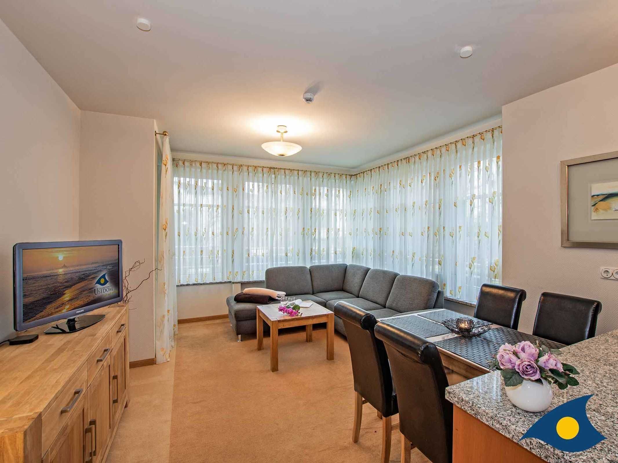 A living room with a sofa, a dining table with chairs, a TV on a wooden cabinet, and a window with curtains.
