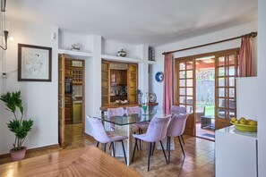 A dining area with a glass table and pink chairs, a kitchen with wooden cabinets, and a view of the garden through large windows. - Beachfront vacation home with 3 bedrooms, air conditioning, and Wi-Fi (Cala D'Or)