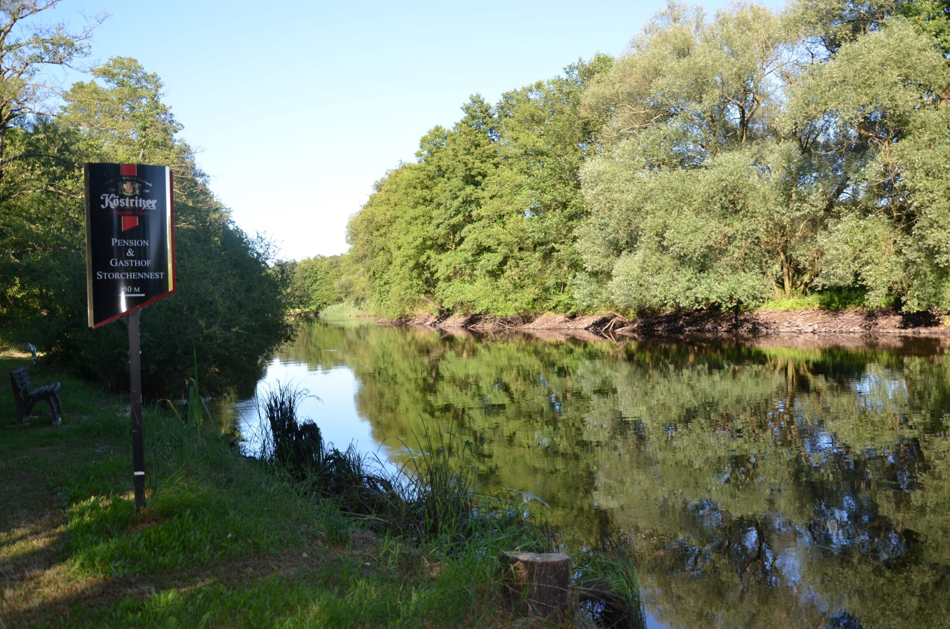 Un cadre paisible au bord de la rivière, avec une enseigne indiquant « Pension Kostringer et auberge Storchennest », et un banc au bord de l’eau.