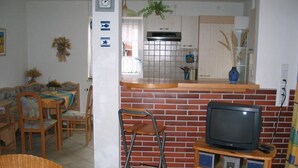 A living room with a TV, a brick fireplace, a dining area with a table and chairs, and a kitchen with cabinets and a window.