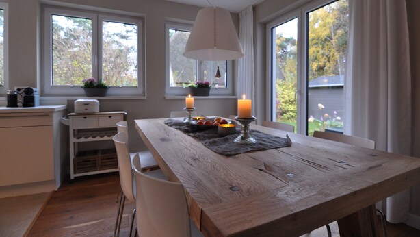 Un coin repas avec une table en bois, des chaises blanches, un luminaire suspendu et une fenêtre donnant sur des arbres.