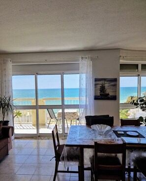 Uma área de refeições com uma mesa posta para dois, grandes janelas com vista para o mar e uma planta em vaso à esquerda.
