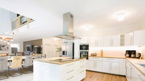 A modern kitchen with white cabinetry, a central island with a built-in stove, and a dining area with a wooden table and chairs. - Villa Thormarcon, Plauer See, 20 Gäste, Sauna+Pool (Malchow)