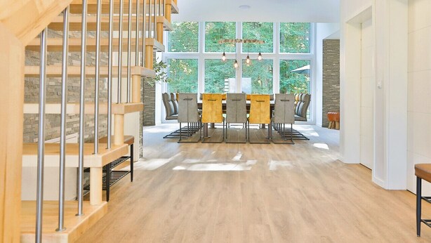 A spacious room with a wooden staircase, a dining area with a table and chairs, and large windows offering a view of greenery outside. - Villa Thormarcon, Plauer See, 20 Gäste, Sauna+Pool (Malchow)