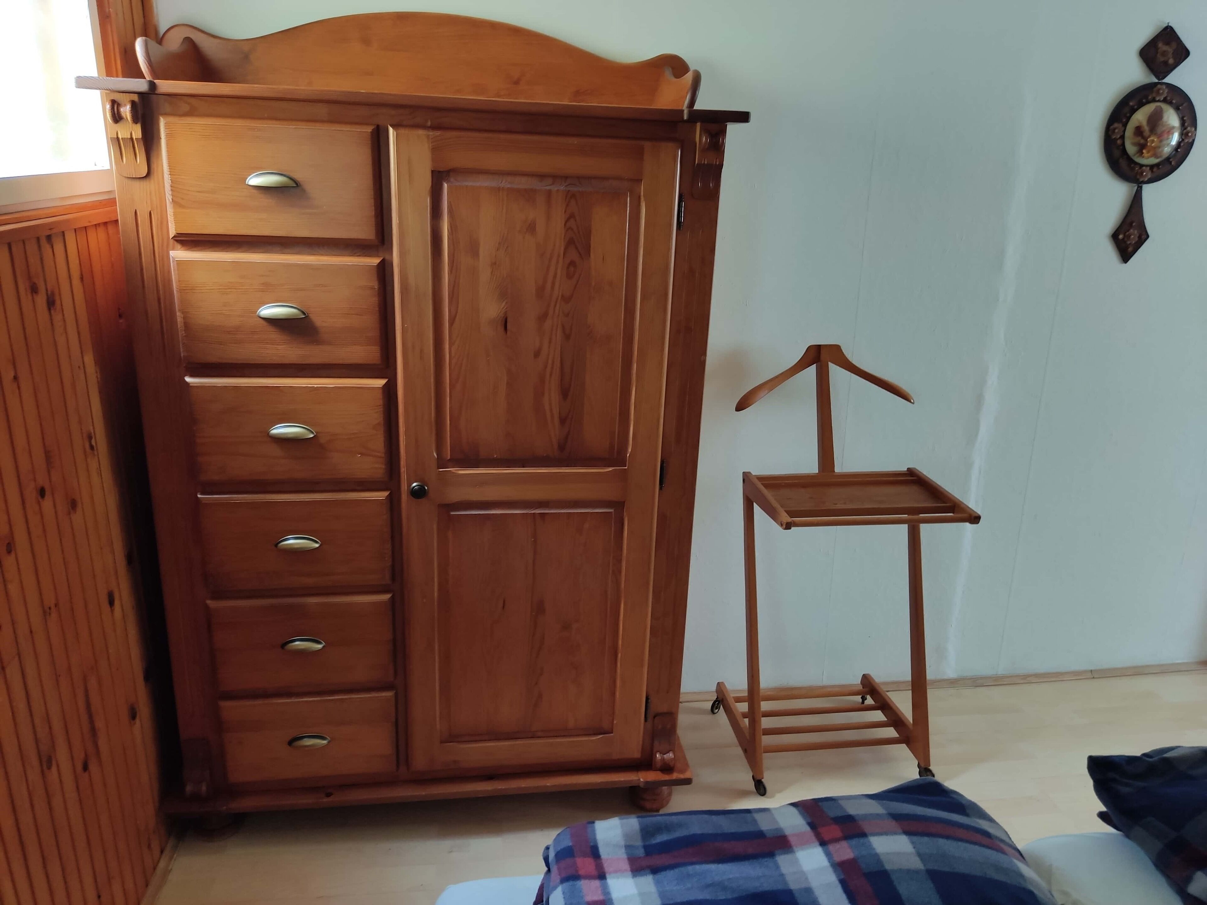 A wooden wardrobe with multiple drawers and a large door, a small wooden table with a hanger, and a plaid blanket on the floor.
