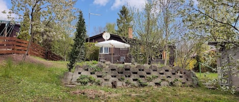 A rustic cabin with a stone wall in front, surrounded by trees and a wooden fence.
