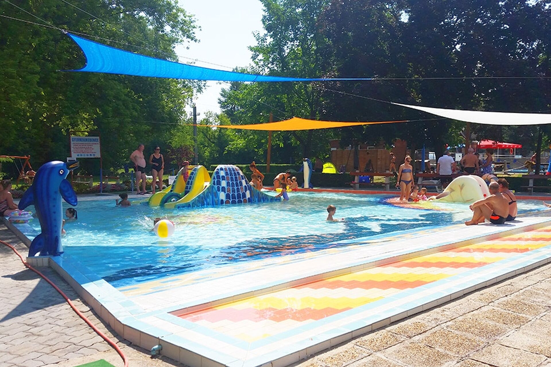 A swimming pool with slides and inflatable toys, surrounded by trees and shaded areas.