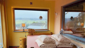 A bedroom with a wooden bed, a window with a view of a green landscape, a small bedside table with a lamp, and a radiator. - How in heaven, colorful house on a hill with stunning views of the Ederta (Hatzfeld)