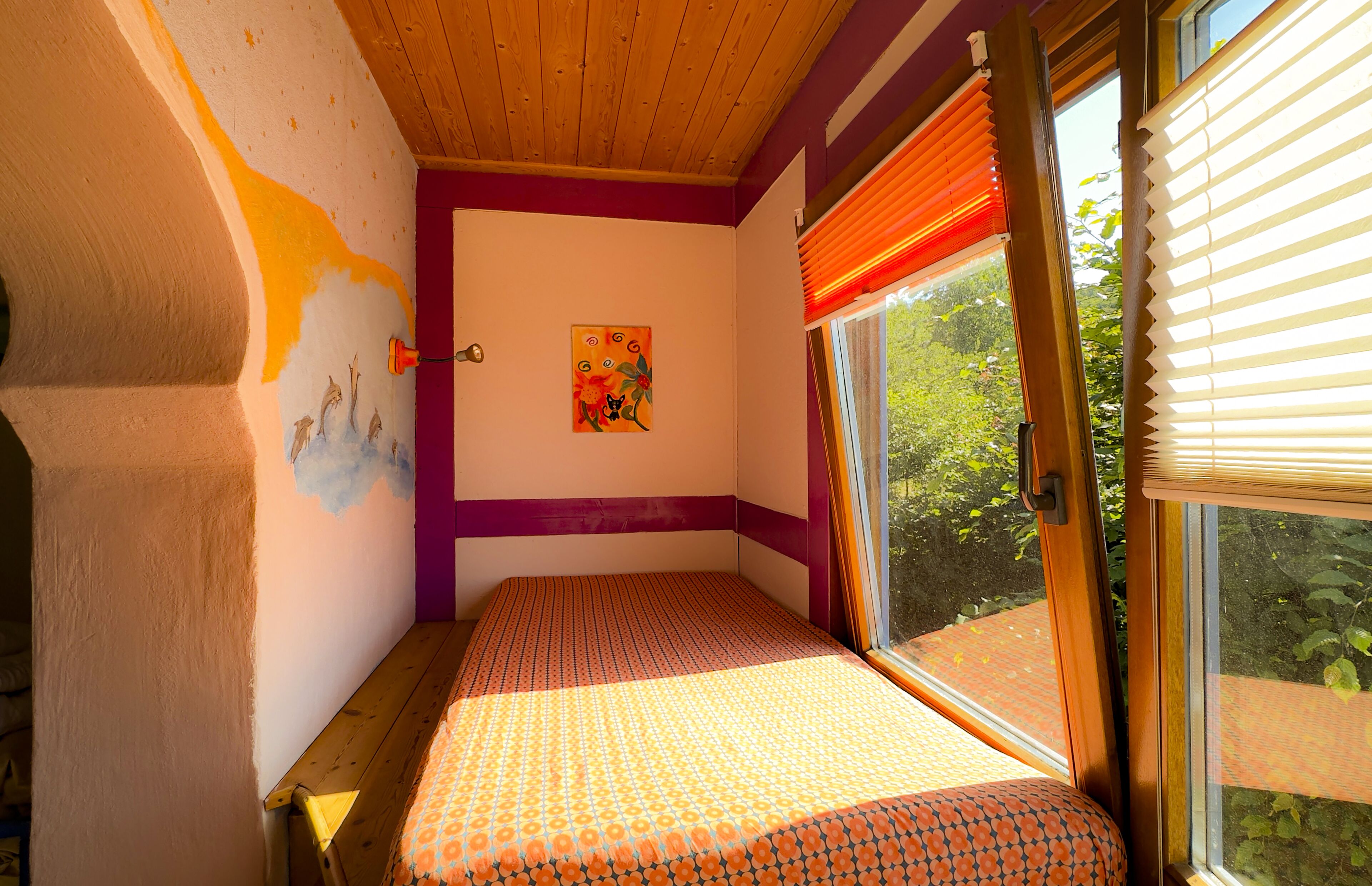 Een kamer met een bed, een raam met jaloezieën, een muur met gedeeltelijk muurschildering en een deur.