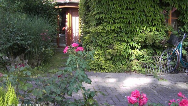 Um jardim com caminho de tijolos, flores cor-de-rosa e uma bicicleta encostada em uma parede.