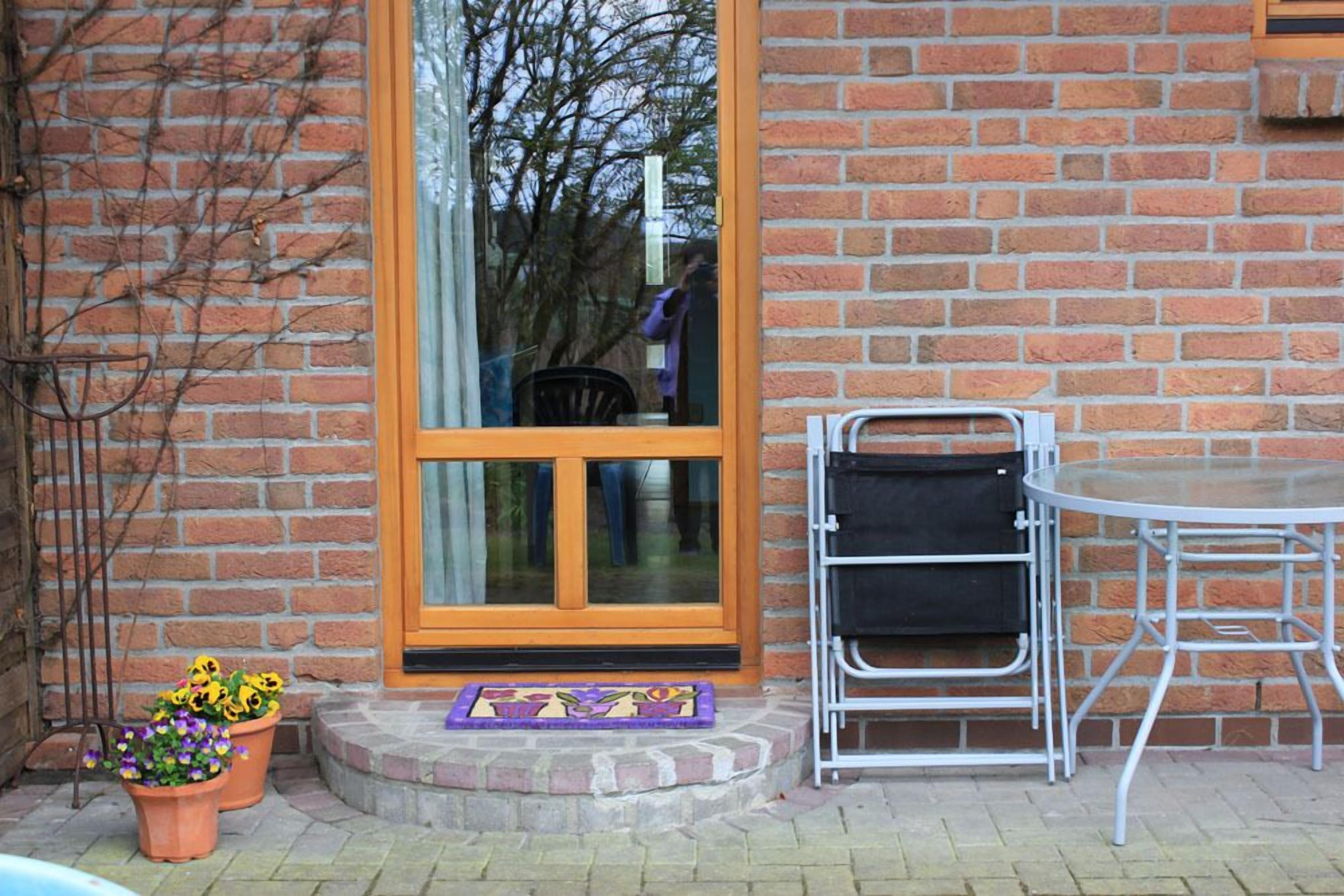 A brick building with a glass door, a small outdoor seating area with a metal table and chairs, and potted plants near the entrance.